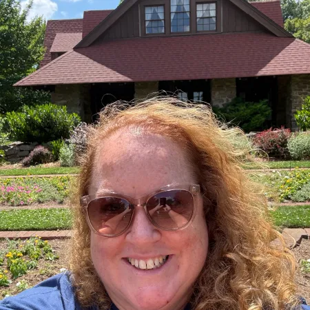 Smiling woman with sunglasses taking selfie in front of a rustic house surrounded by greenery on a sunny day