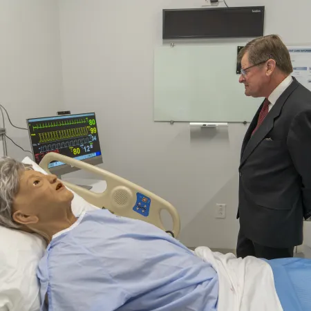 Man in suit observing medical mannequin in hospital bed with vital signs monitor in clinical simulation room.