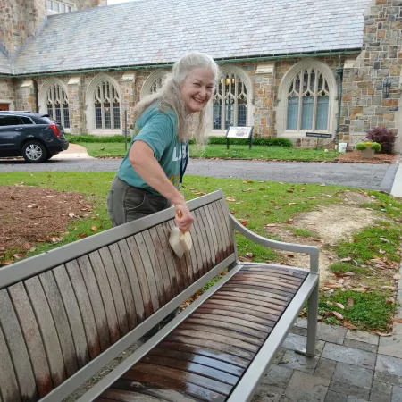 Smiling elderly woman cleaning an outdoor wooden bench in a courtyard with stone building and parked car in background