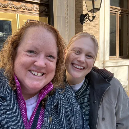 Two smiling women wearing coats standing outside a building with yellow doors and brick walls
