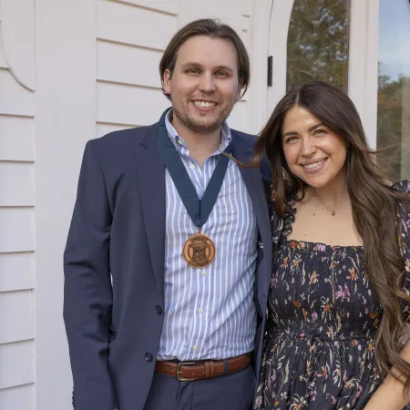 Smiling young man wearing a medal and young woman in floral dress stand outside a white building with a glass door.