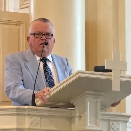 Man in light gray suit speaking at ornate white church pulpit with microphone and cross symbol.