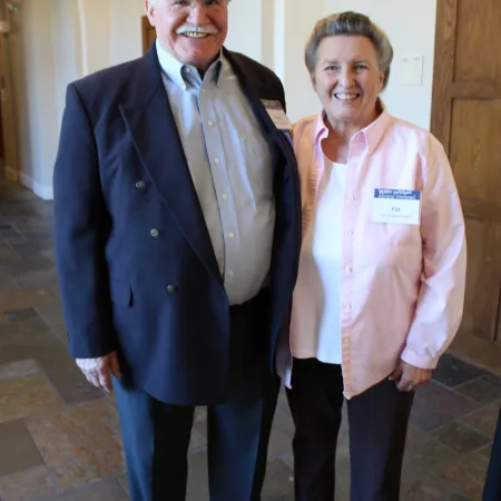Two smiling people posing indoors, one in a dark suit and one in a light pink shirt, standing on tiled floor.