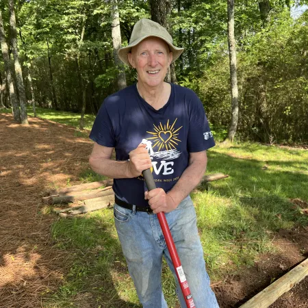 Smiling elderly man wearing hat and gloves holds a shovel while gardening in a sunlit wooded area.