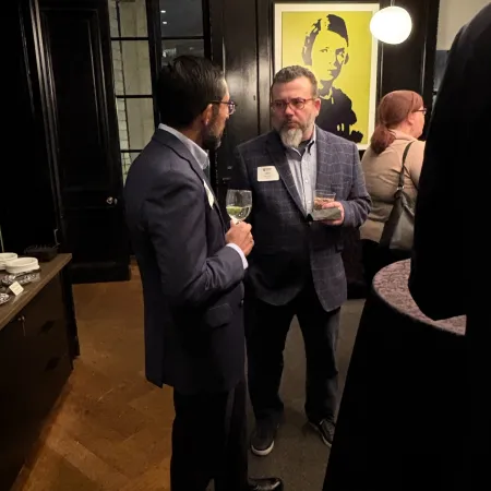 Two men engaged in conversation at an indoor event with drinks in hand and artwork on the wall.