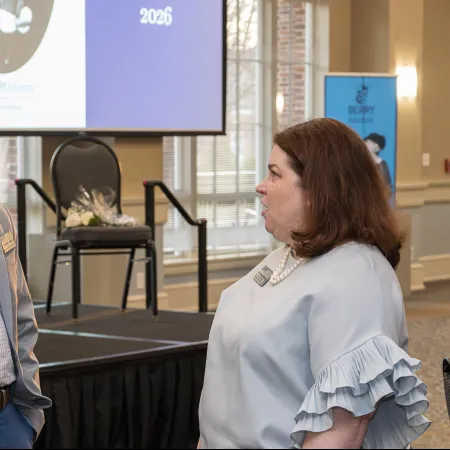 Two professionals engaged in conversation near a stage at a conference with presentation screen in background.