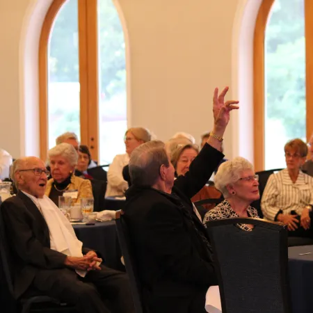 Seniors and adults engaged in discussion at a formal event with tables, drinks, and large arched windows.