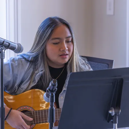 Young woman playing acoustic guitar and singing with microphones and sheet music stand indoors.