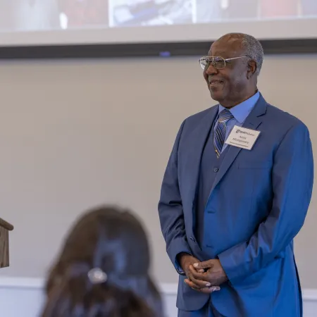 Woman speaking at podium during event with man in blue suit standing to the side, audience visible.