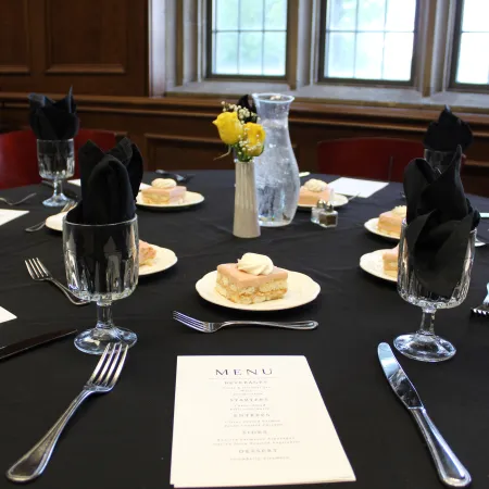 Elegant dining table with black tablecloth, black napkins, glassware, menus, and small dessert plates in a wood-paneled room.