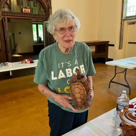 Elderly woman in green Labor Day shirt holding a carved wooden turtle inside a community hall.