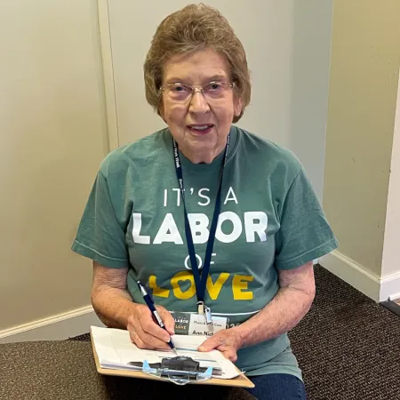 Elderly woman in glasses wearing a green shirt with It's a Labor of Love writing, writing on a clipboard indoors.