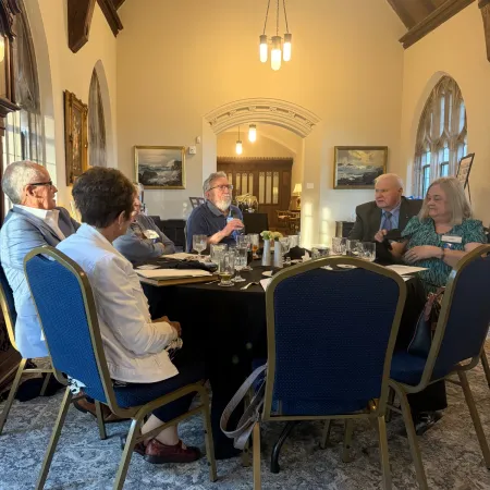 Group of older adults having a discussion around a table in a warmly lit room with blue chairs and arched windows.
