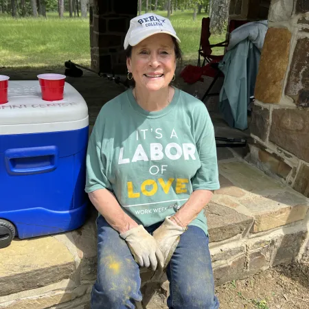 Smiling woman in work gloves and labor of love shirt sitting on stone ledge next to blue cooler with red cups.