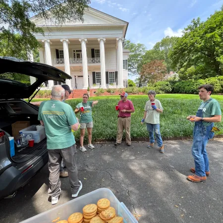 Group of people socializing outdoors with drinks and snacks near a historic white columned mansion under blue sky.