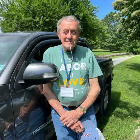 Elderly man in green T-shirt standing by black Tacoma truck on sunny day with trees and grass landscape.