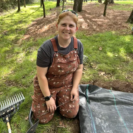 Smiling woman in floral overalls kneeling on grass next to gardening tools and tarp outdoors in sunlight.