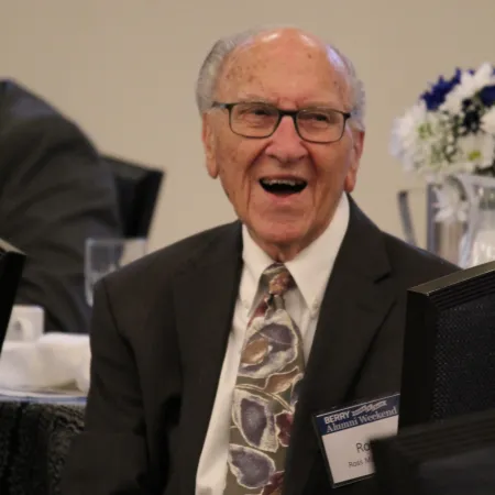 Elderly man in suit and floral tie speaking at an event with name badge and flowers in background