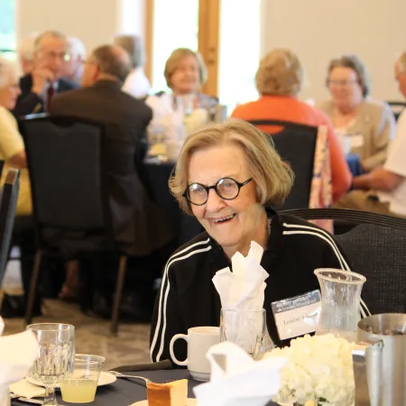 Two elderly women enjoy conversation at a social event with drinks and table settings in a cozy room.