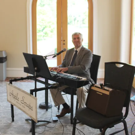 Man in suit playing keyboard and singing indoors near large windows with greenery outside.