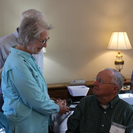 Two elderly people engaging in a conversation indoors with a lit lamp in the background.