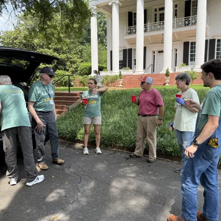 Group of six people chatting outside a white colonial house, some holding red cups, one unloading a car trunk.