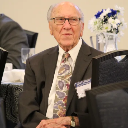 Elderly man in glasses wearing a suit and floral tie seated at a table with flower centerpiece in background