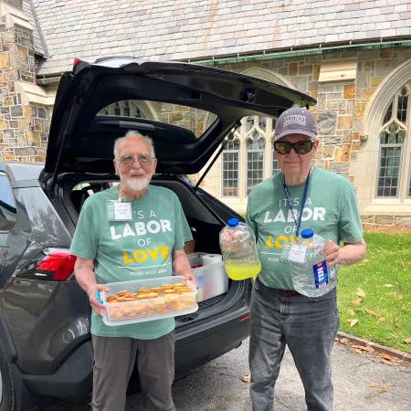 Two elderly men wearing matching green Labor of Love shirts stand by a car trunk holding snacks and drinks outdoors.