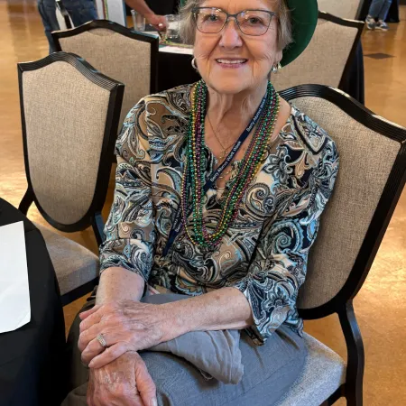 Elderly woman wearing a green hat, glasses, patterned blouse, and Mardi Gras beads sitting on a chair indoors.