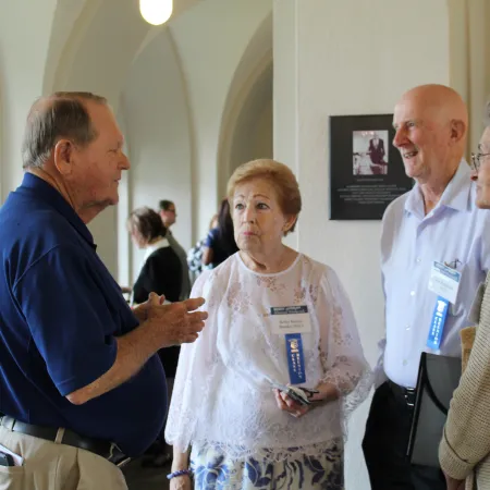 Four elderly people engaging in conversation at an indoor event with name tags and ribbons.