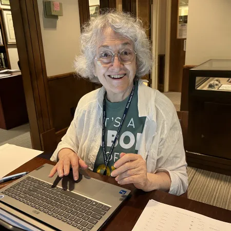 Smiling elderly woman with curly white hair using a laptop in a cozy library setting with books and documents.