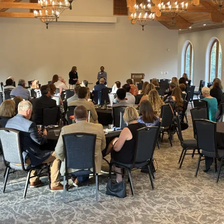 Audience seated at round tables listening to two presenters in a wood-ceiling conference room with large windows.