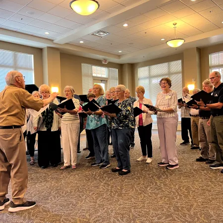 Senior choir group rehearsing indoors led by conductor holding sheet music in a well-lit room.