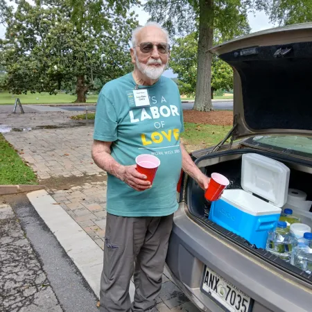 Elderly man wearing sunglasses and Labor of Love shirt holding red cups by a car trunk with water bottles and cooler.