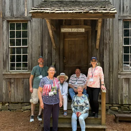 Group of six adults posing in front of a rustic wooden cabin with a porch and sign reading Museum East Church.