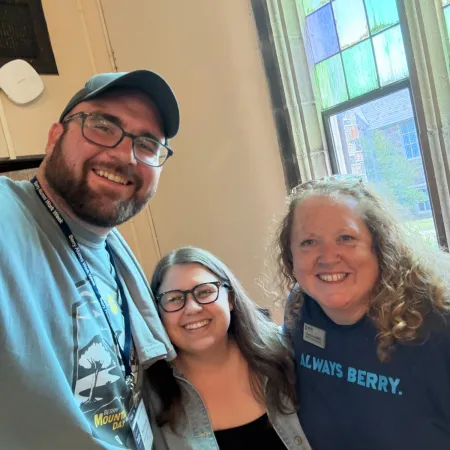 Three smiling people posing indoors near a window with stained glass, wearing casual clothing and glasses.