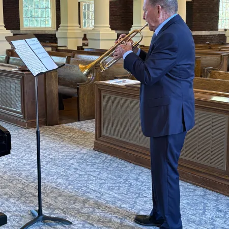 Man in blue suit playing trumpet in a room with wooden pews and carpeted floor beside a music stand.