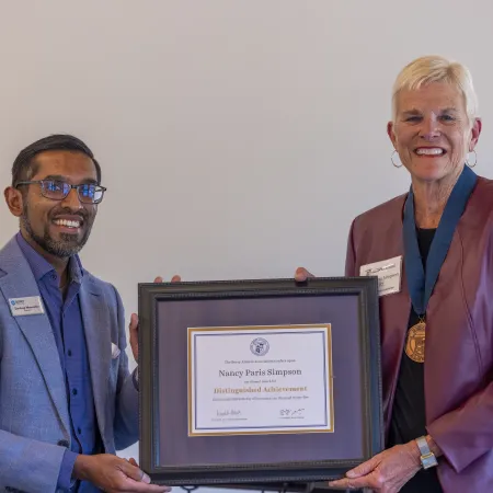 Two people smiling and holding a framed Distinguished Achievement certificate in a formal setting