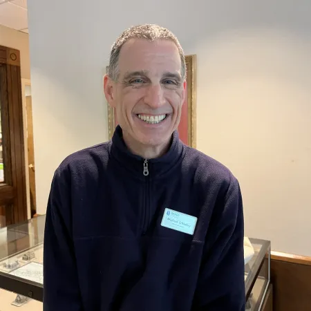 Smiling man wearing a navy zip-up sweater with a name tag standing indoors near a glass display case.