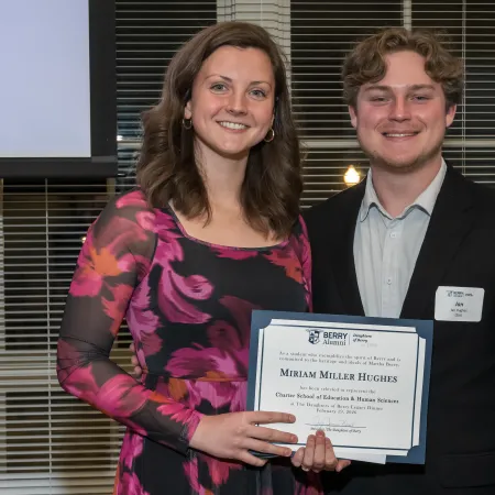 Two smiling individuals holding a certificate of recognition at a formal award event indoors.