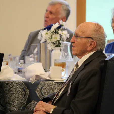 Elderly men in suits sitting at a formal event table with flowers and glassware, attentively listening.