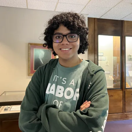 Young person with curly hair and glasses smiling indoors wearing a green hoodie and graphic t-shirt.