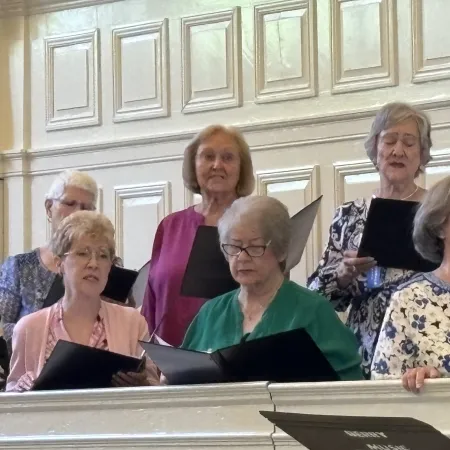 Senior women singing in a choir inside a church with hymnals and formal attire.