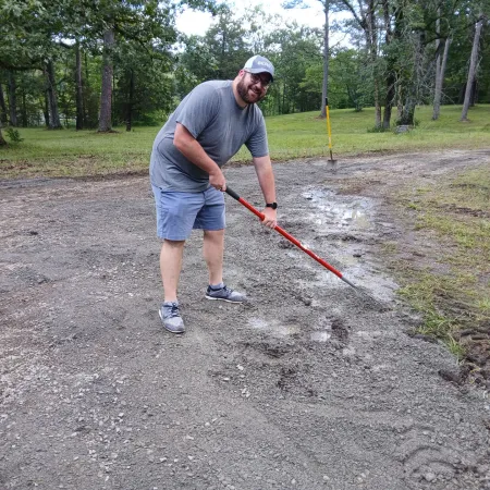 Man in casual clothes using a red rake on a gravel path next to grassy area with trees in background.