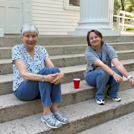 Two women sitting on concrete steps outside a white building, smiling and engaging in conversation.