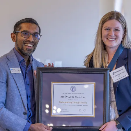Two smiling professionals holding a framed Outstanding Young Alumni award certificate inside a bright room.