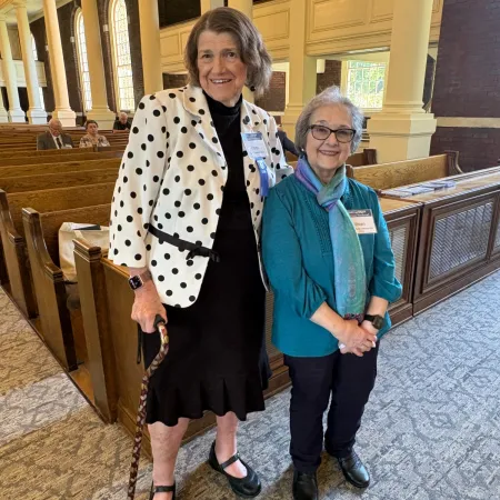 Two older women standing inside a carpeted room with wooden pews and large windows, one holding a cane.