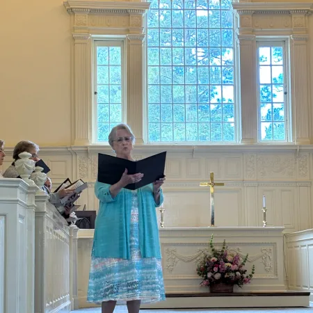 Woman in blue dress singing in a church with large arched windows and white walls behind her