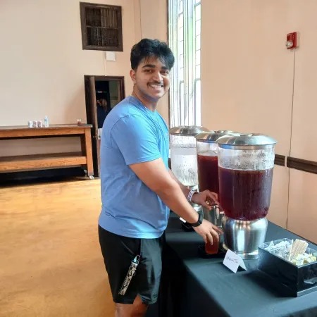 Smiling young man in blue shirt pouring drink from large beverage dispenser at indoor event.
