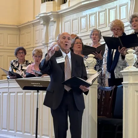 Senior man in suit conducting choir of elderly women singing in elegant room with organ pipes.